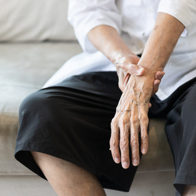 An elderly person with visible hand veins holds their own wrist to steady a resting tremor, sitting next to a cane -- health tech coverage from STAT