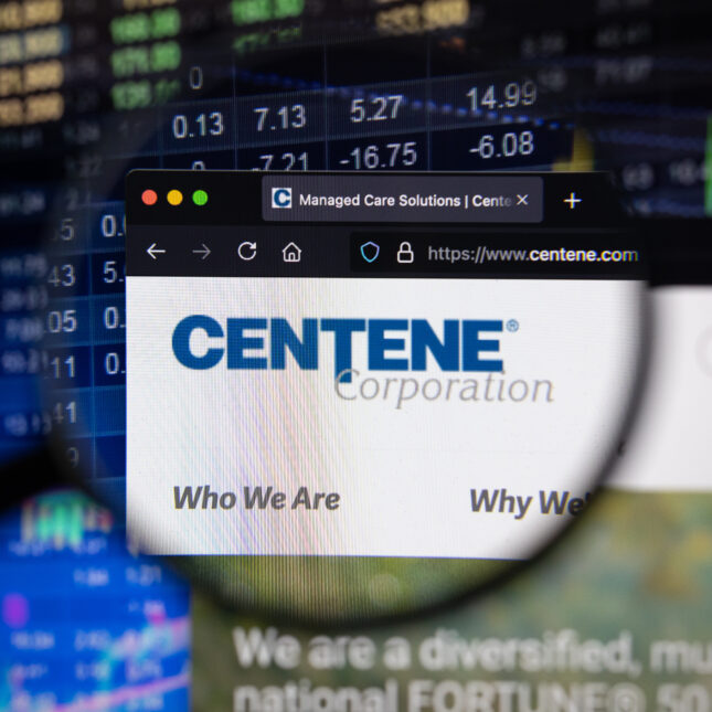 Centene company logo on a website with blurry stock market developments in the background, seen on a computer screen through a magnifying glass. -- health policy coverage from STAT