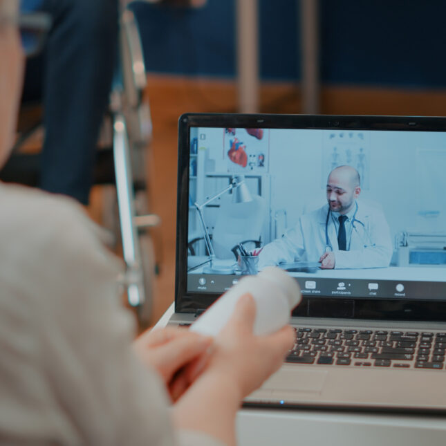 A senior, holding a bottle of medicine, on videocall with a doctor at home — coverage from STAT