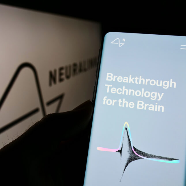 A smartphone displaying Neuralink's website with the text "Breakthrough Technology for the Brain" is held in front of a larger screen showing the Neuralink logo -- health tech coverage from STAT