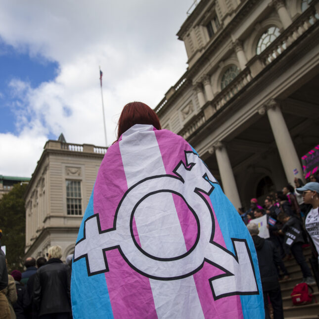 A rally participant stands in front of the New York City Hall, with a transgender flag with a transgender symbol on it wrapped around them -- coverage from STAT