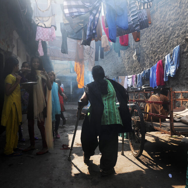 File photograph of a polio patient in Kolkata India walking between two stones walls with walking aids. colorful clothing hangs above her on a clothes line.