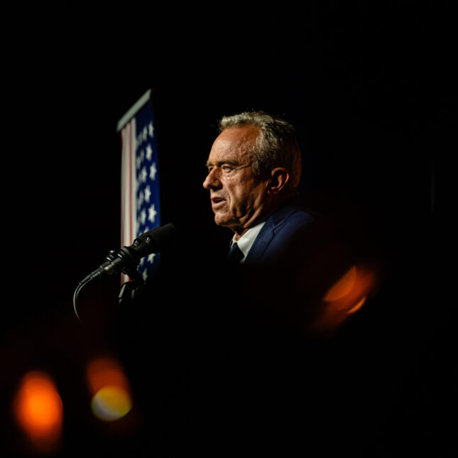 A spotlight illuminates the face of Robert F. Kennedy Jr. as he speaks and an American flag further next to him -- politics coverage from STAT