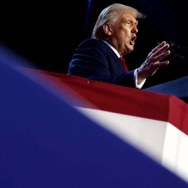 Trump speaks and gestures is in focus behind the blurry foreground of red, white, and mainly blue stage decoration -- coverage from STAT